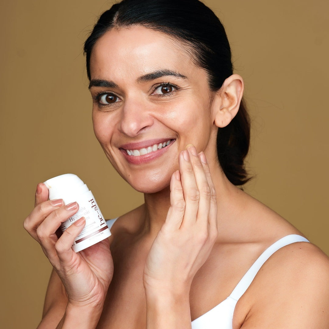Image of a smiling woman holding the He-shi Overnight Hyaluronic Tan Balm in her right hand and touching her left hand to her face